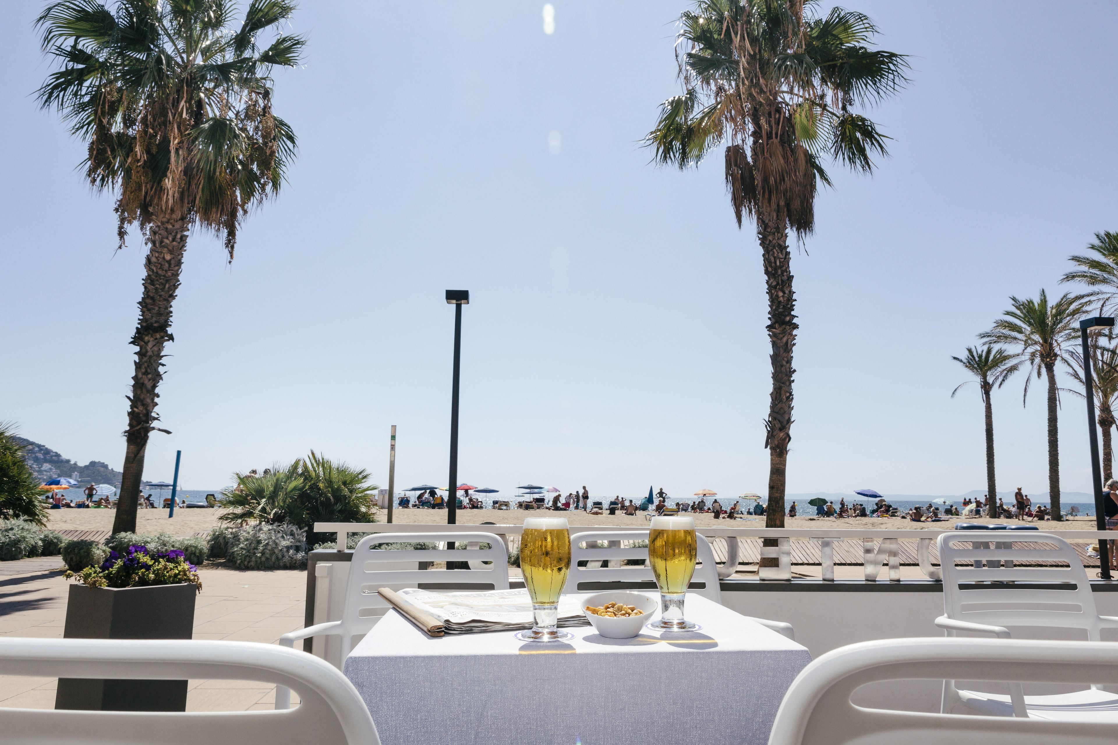 Una mesa con dos vasos de una bebida y un bol de nueces, con vista a una playa con palmeras y gente a lo lejos.