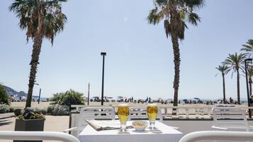 Una mesa con dos vasos de una bebida y un bol de nueces, con vista a una playa con palmeras y gente a lo lejos.