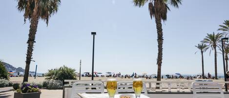 Una mesa con dos vasos de una bebida y un bol de nueces, con vista a una playa con palmeras y gente a lo lejos.