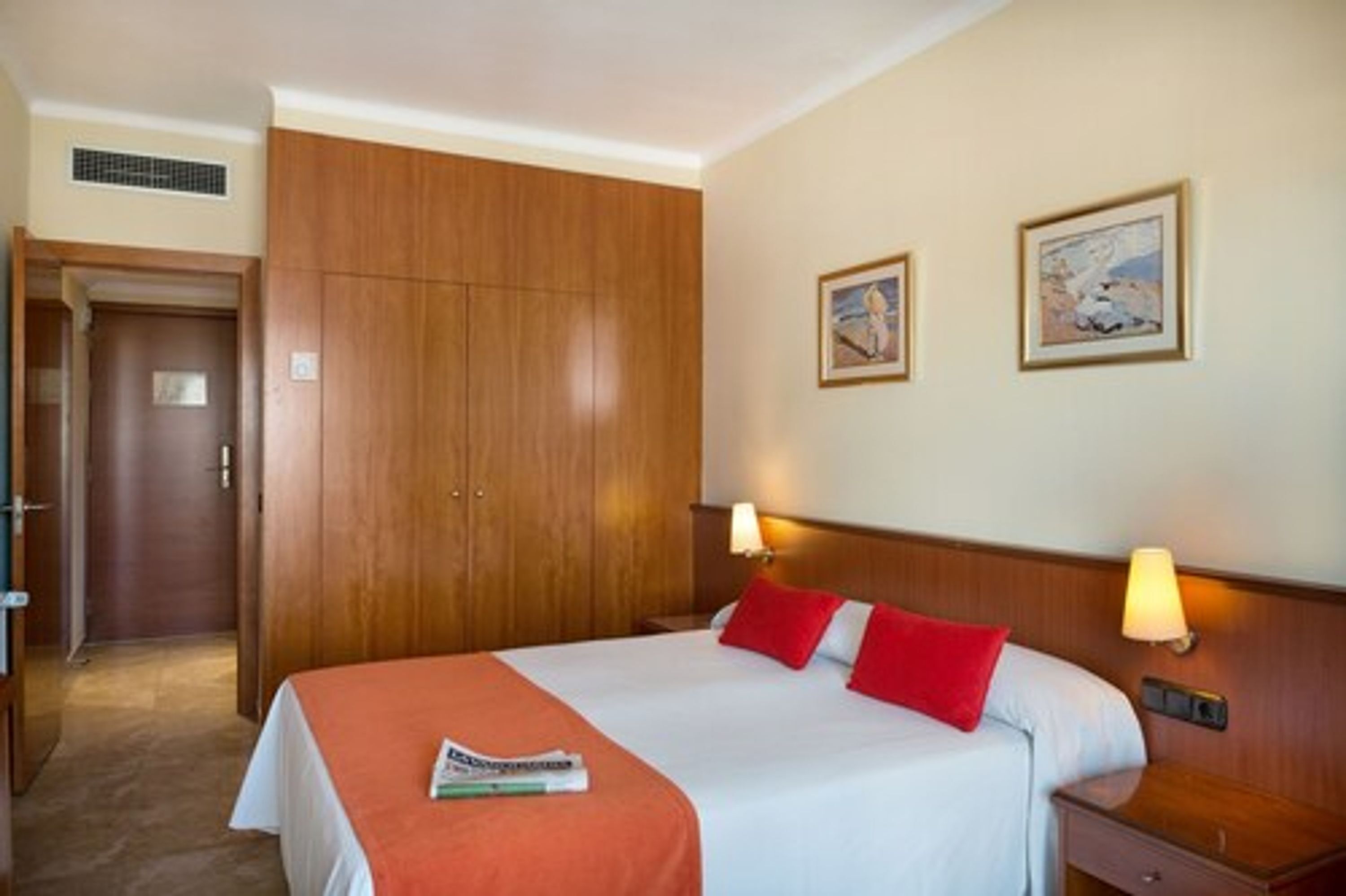 Una habitación de hotel con una cama grande, dos lámparas de noche, un armario de madera y dos cuadros en la pared.