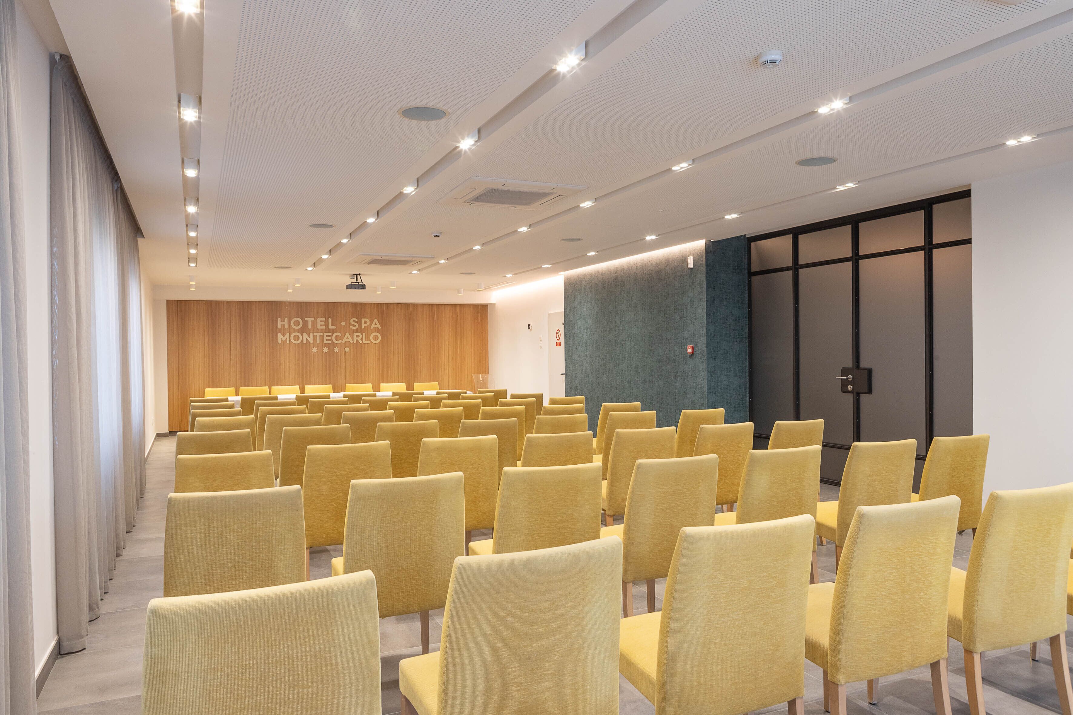 A conference room with rows of yellow chairs, a wooden panel wall with the text "HOTEL SPA MONTECARLO," and a dark-colored door.