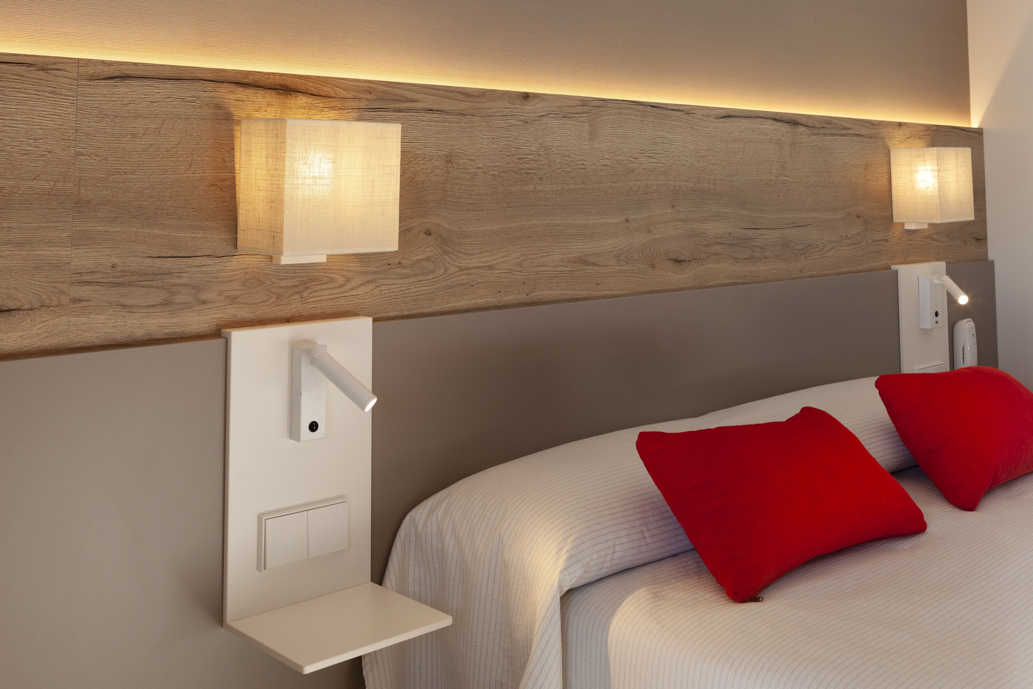 A modern hotel room with a wooden headboard, two bedside lamps, a white bedside table, and two red pillows.