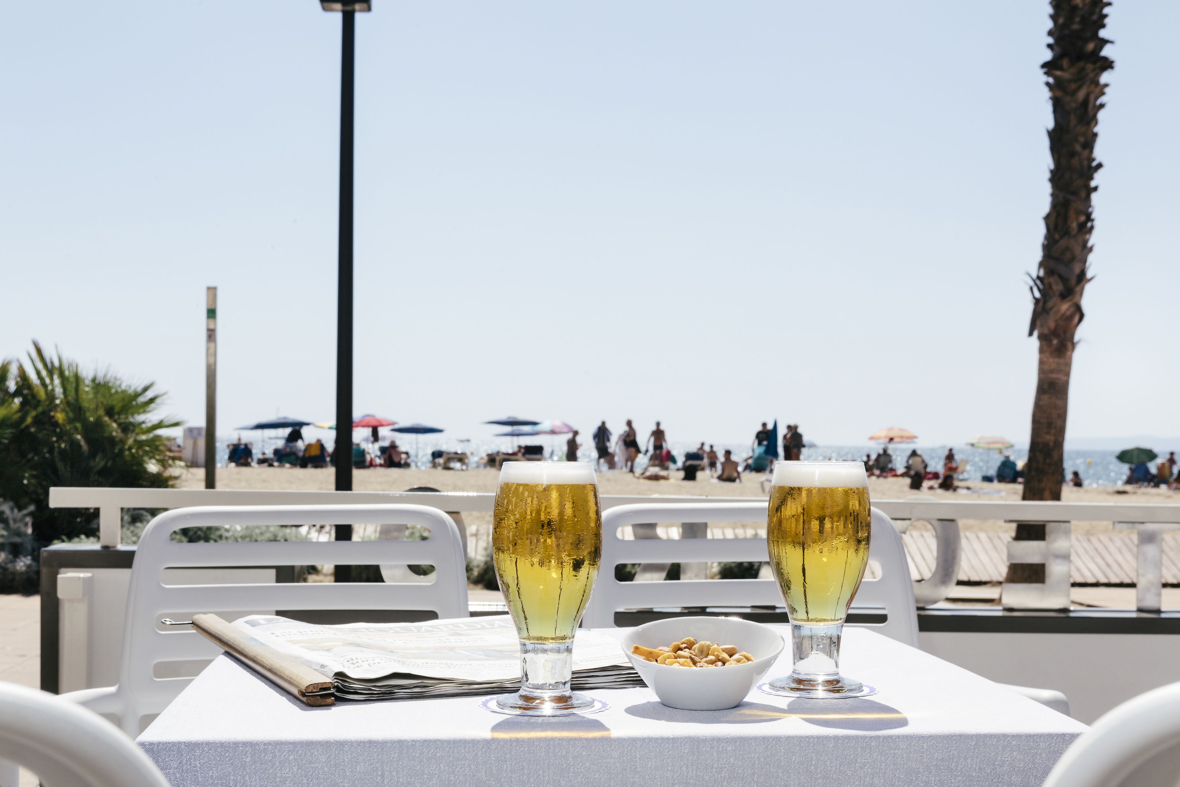 Une table avec deux verres de bière et un bol de grignotages, donnant sur une plage avec des parasols et des gens.