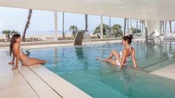 An indoor swimming pool with clear views of the beach and palm trees.