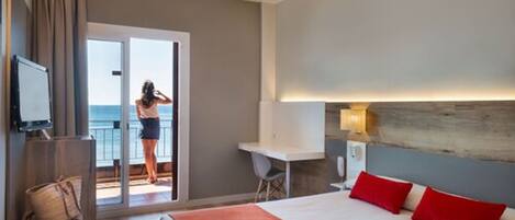 A hotel room with a bed, a desk, a chair, a television, and a balcony with a view of the sea.