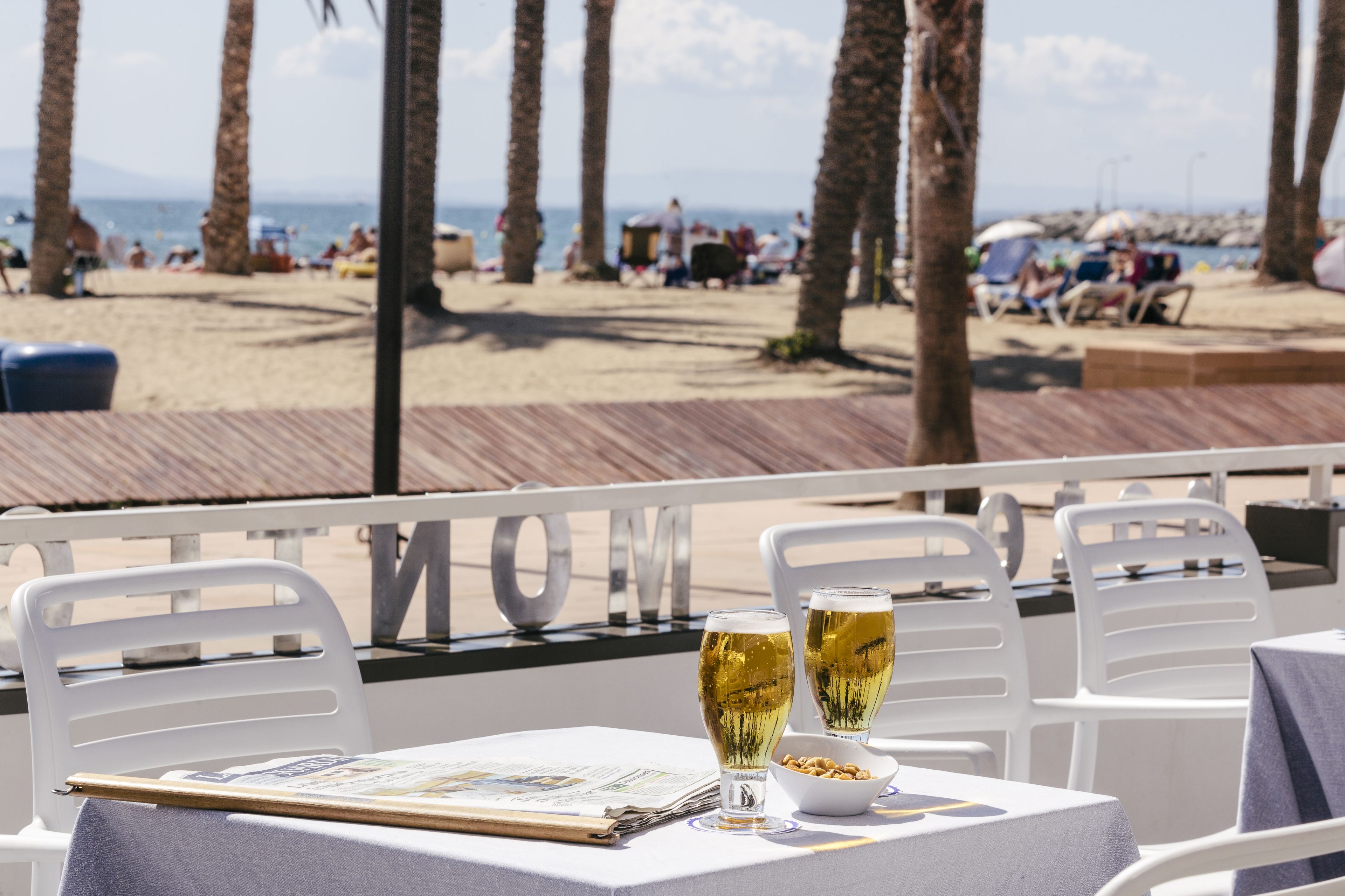 Una mesa con dos vasos de cerveza, un menú y un bol de frutos secos en un restaurante frente al mar, con vistas a la playa y palmeras.