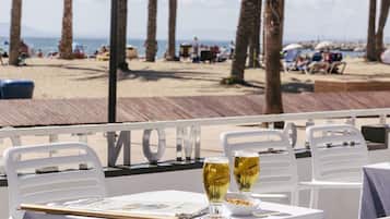 Una mesa con dos vasos de cerveza, un menú y un bol de frutos secos en un restaurante frente al mar, con vistas a la playa y palmeras.