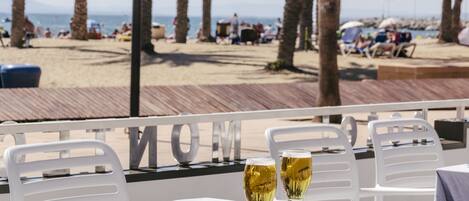 A table with two glasses of beer, a menu, and a bowl of nuts at a beachside restaurant with a view of the beach and palm trees.