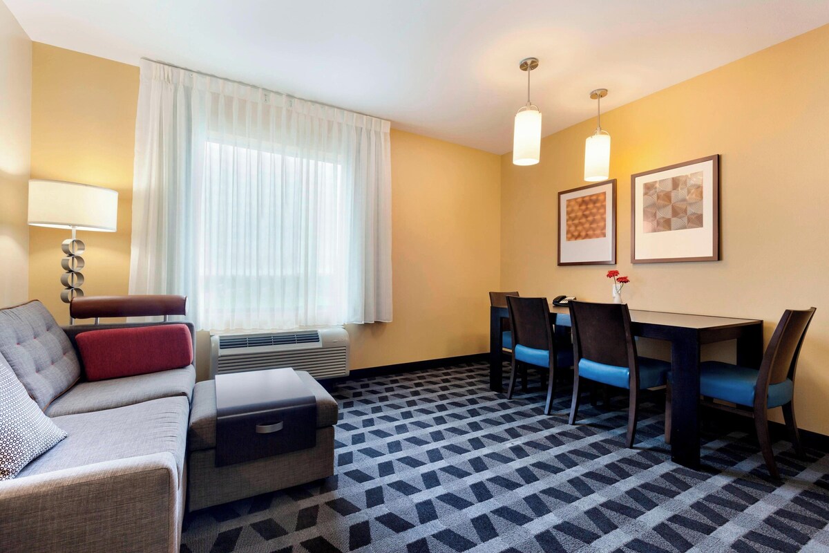 Une chambre d’hôtel avec un canapé, une petite table, une table à manger avec des chaises, une lampe et deux tableaux encadrés au mur.