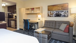 Habitación de hotel con cama, sofá, escritorio con silla, kitchenette y obras de arte en la pared.