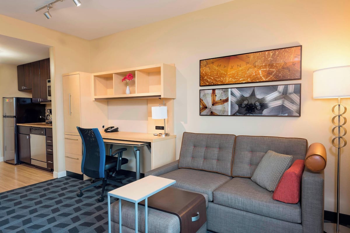 Una sala de estar moderna con un sofá gris, una mesita y un escritorio con una silla. Se observa una zona de cocina con armarios y un refrigerador.