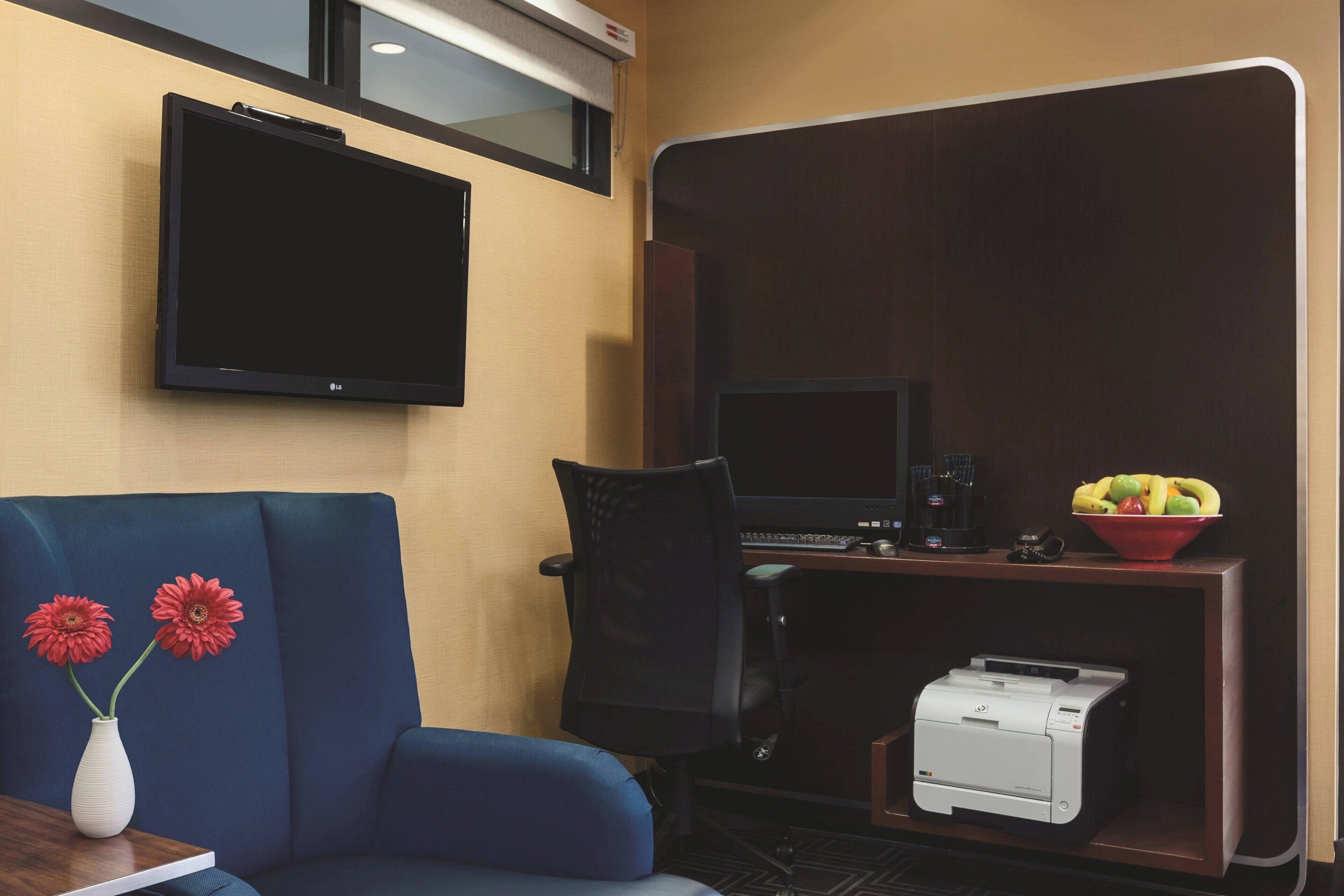 A hotel room with a flat-screen TV mounted on the wall, a desk with a computer and a printer, a blue sofa, and a vase with flowers.