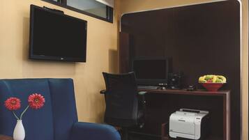 A hotel room with a flat-screen TV mounted on the wall, a desk with a computer and a printer, a blue sofa, and a vase with flowers.
