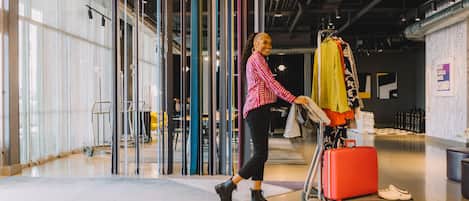 Una persona empuja un carrito de equipaje con una maleta roja y una muda de ropa amarilla en un espacio interior moderno con ventanas grandes y estructura de techo visible.