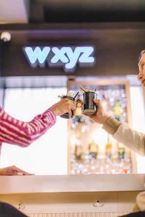 Two people toasting with drinks at a bar with a 'WXYZ' sign in the background. - Aloft by Marriott Frisco (Frisco)