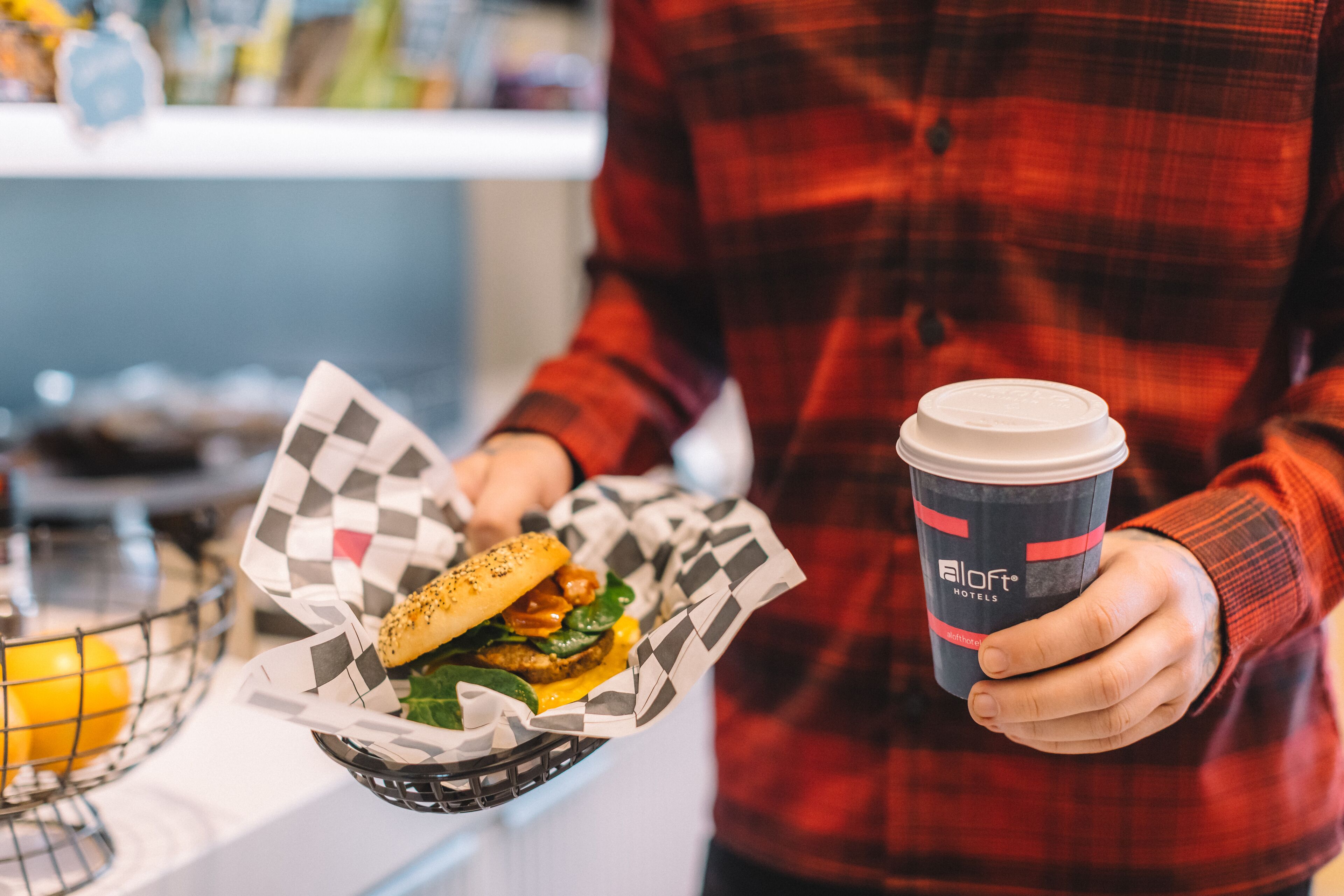 A person holding a burger wrapped in checkered paper and a cup with a lid.