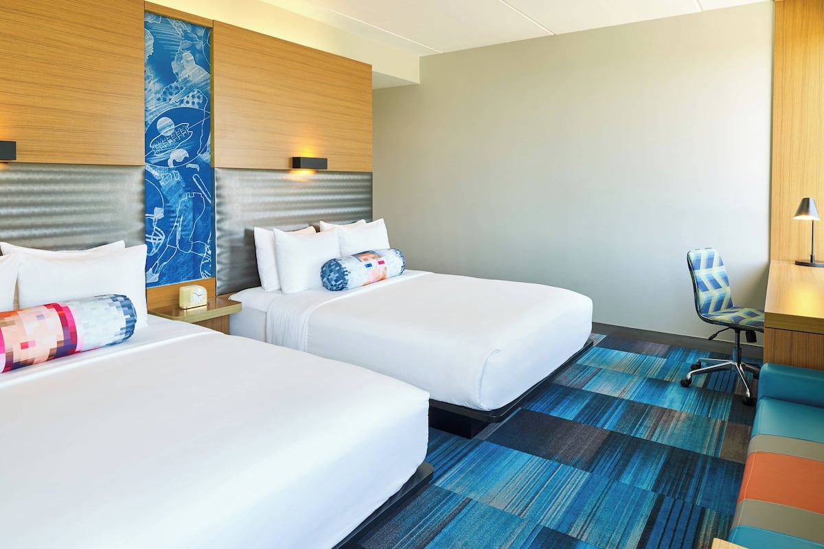 A hotel room with two beds, a blue patterned wall, a blue carpet, and a chair.