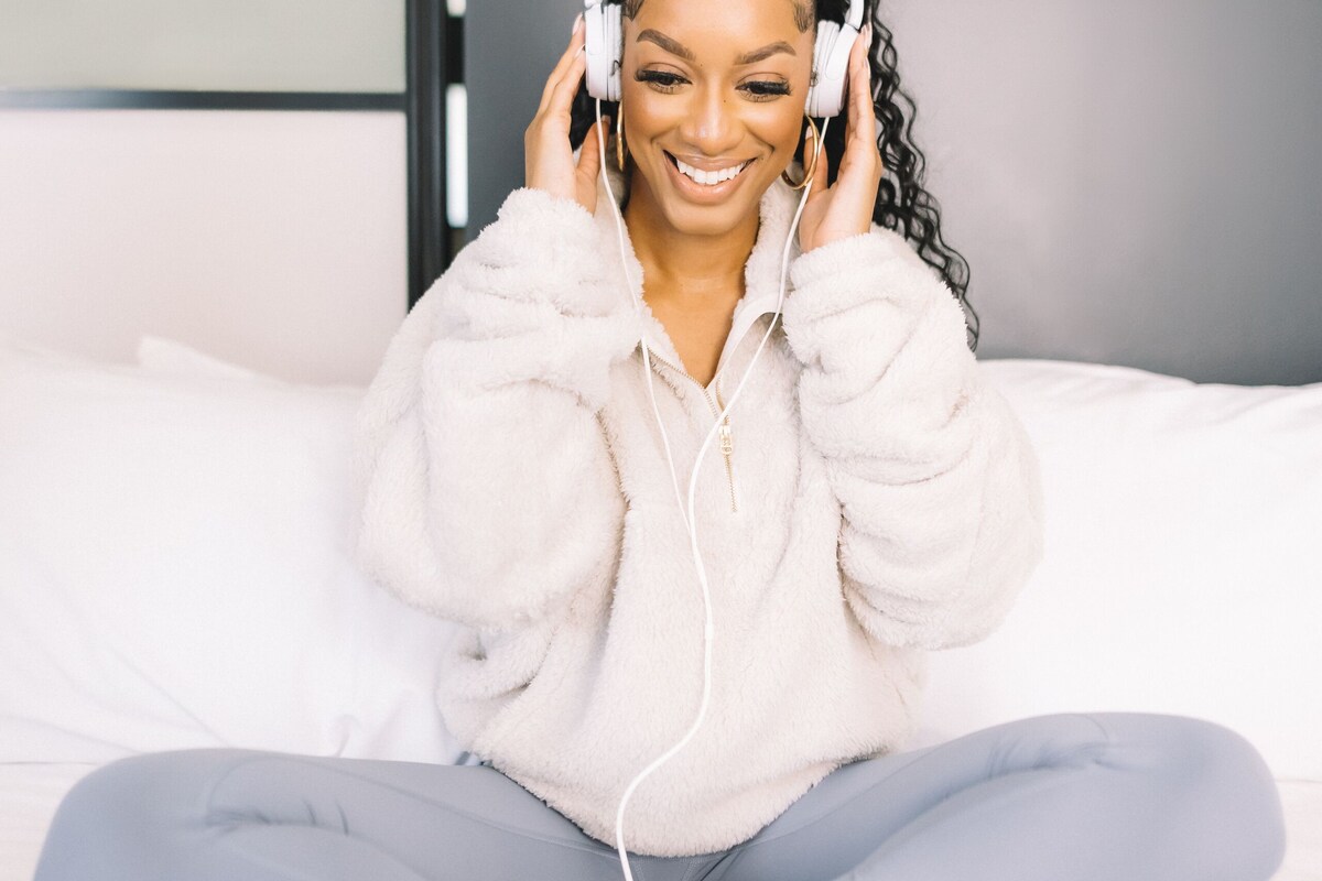 A person sitting on a bed with a laptop, wearing headphones, and holding a phone.