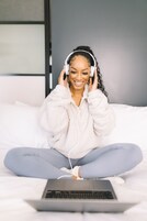 Una persona sentada en una cama con una computadora portátil, audífonos y sosteniendo un teléfono.