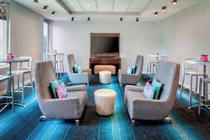 Uma área de estar moderna com poltronas cinzas, um banquinho central e uma TV de tela plana fixada em uma parede de madeira.