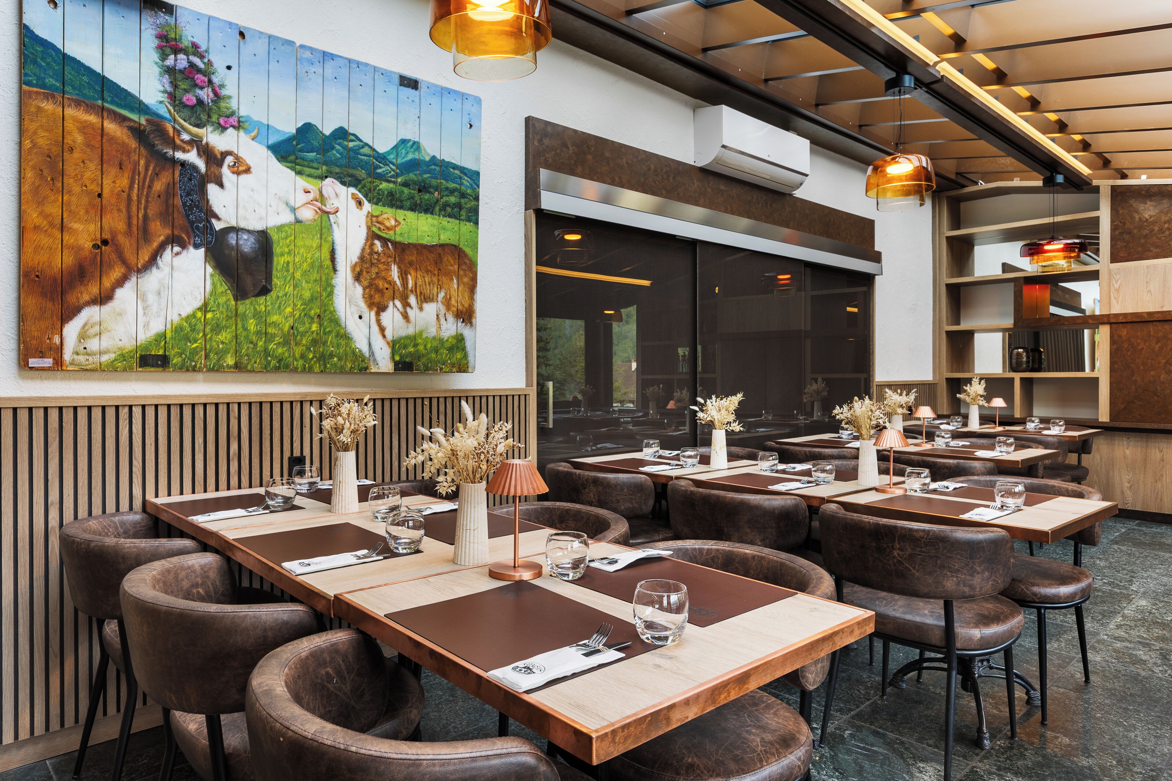 Un restaurante con mesas de madera y sillas de cuero, un mural grande de vacas e iluminación colgante.
