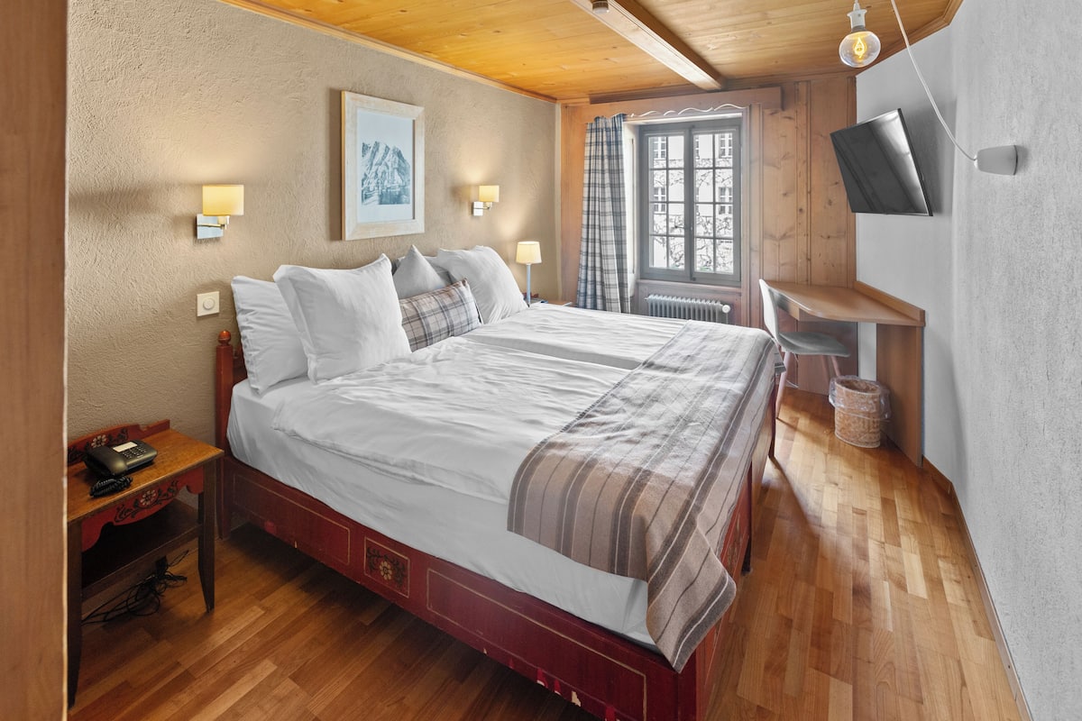A bedroom with a wooden bed, bedside lamps, a mounted TV, and a window with curtains.