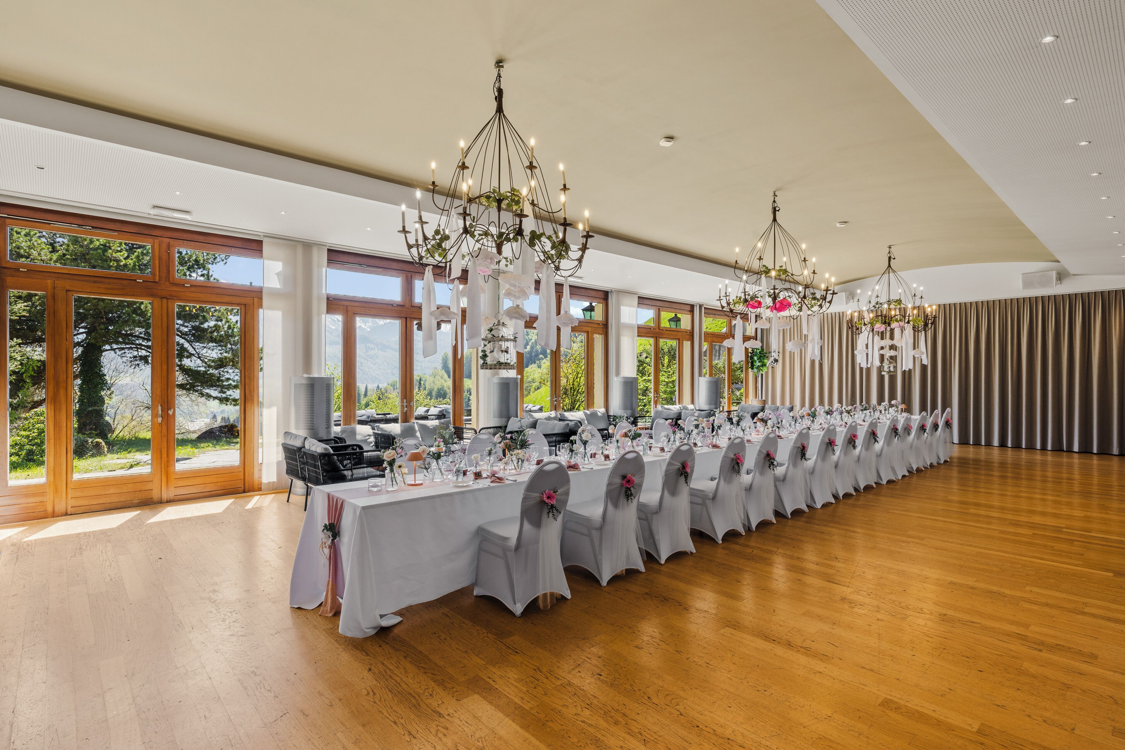 Una larga mesa de banquete con manteles blancos y sillas, candelabros y ventanas amplias con vistas a zonas verdes.