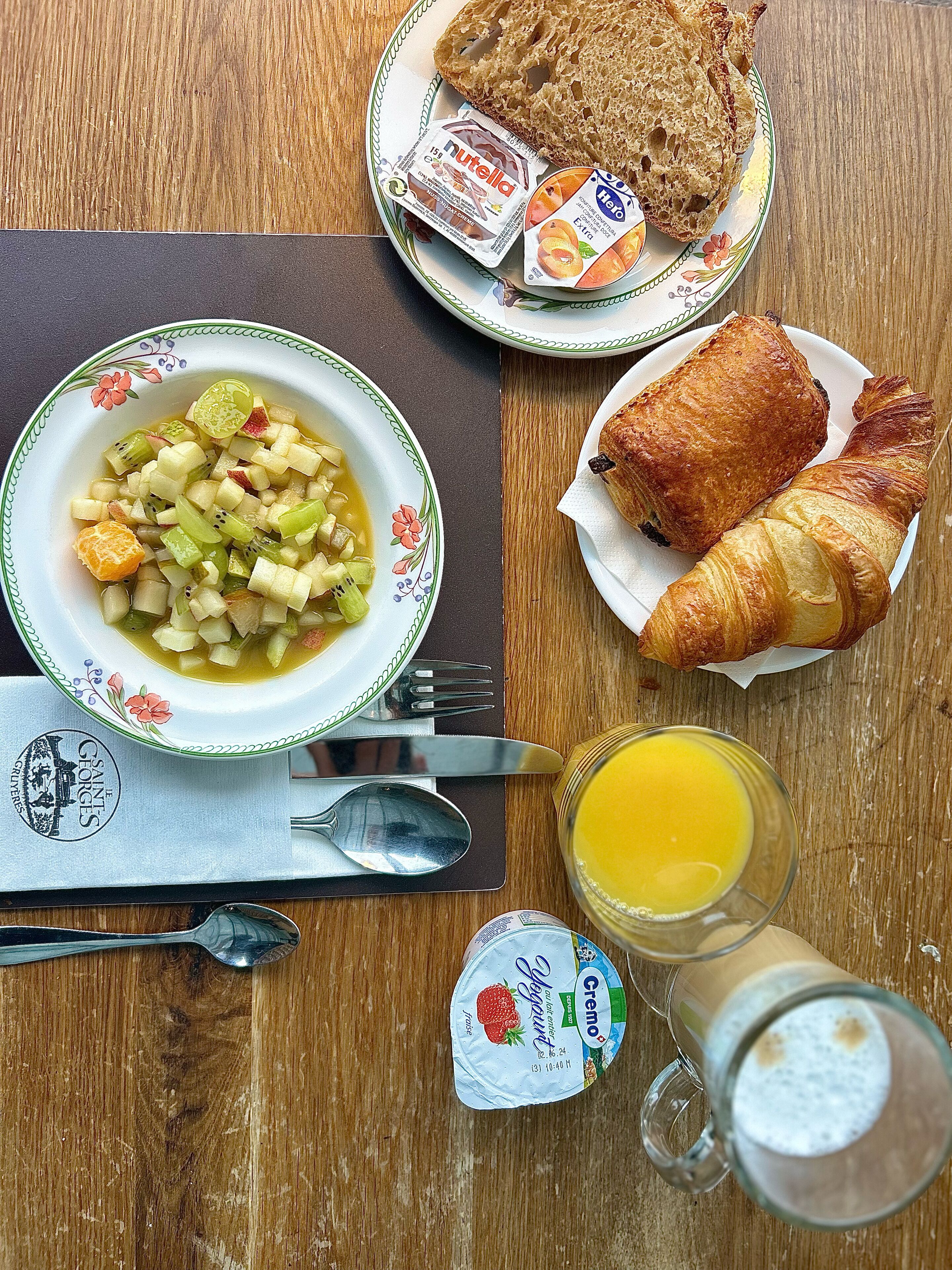 Esmorzar amb un bol de amanida de fruita, croissants, Nutella, iogurt i suc d'aranja damunt una taula de fusta.