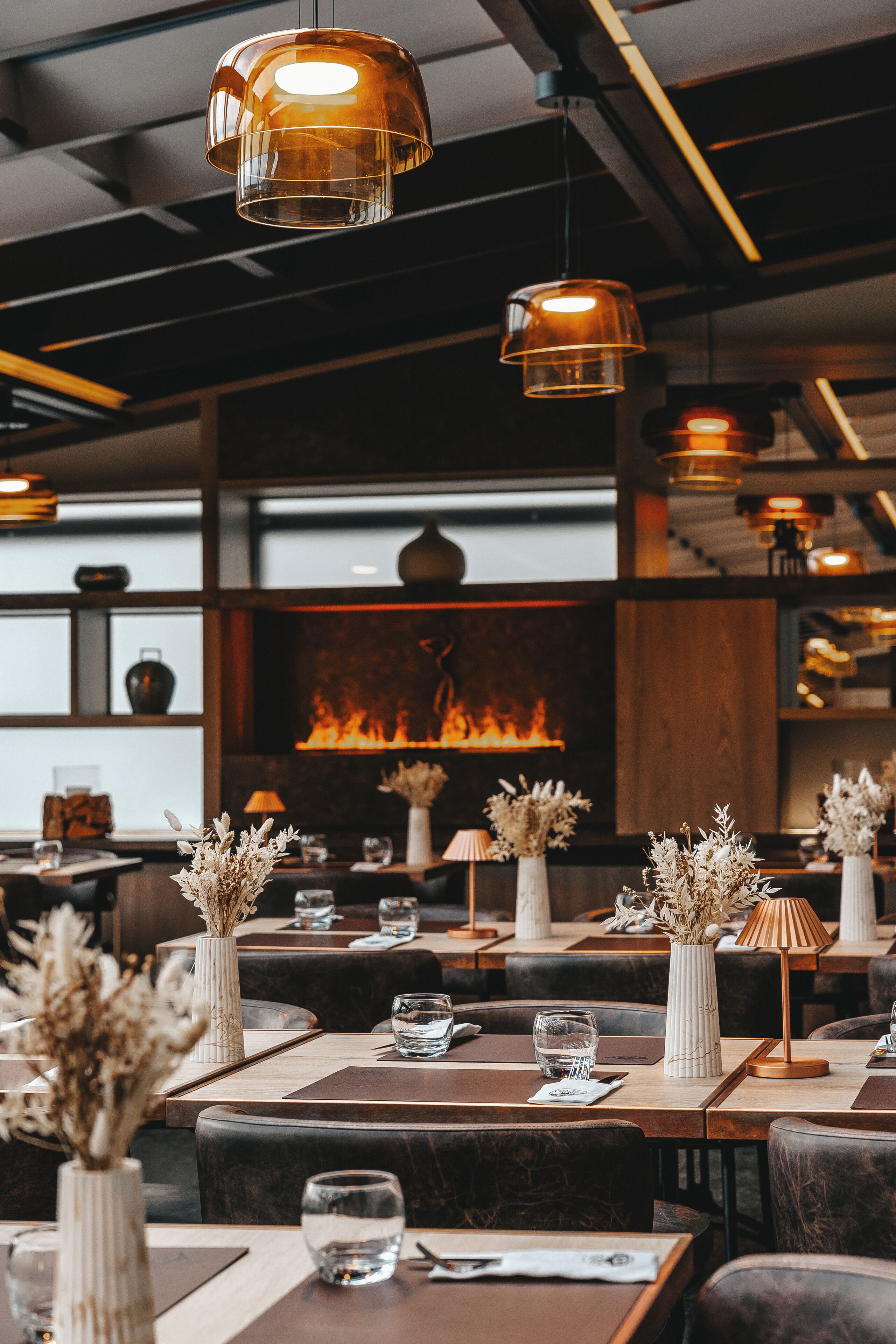 A restaurant interior with tables set for dining, a fireplace in the background, and hanging pendant lights.