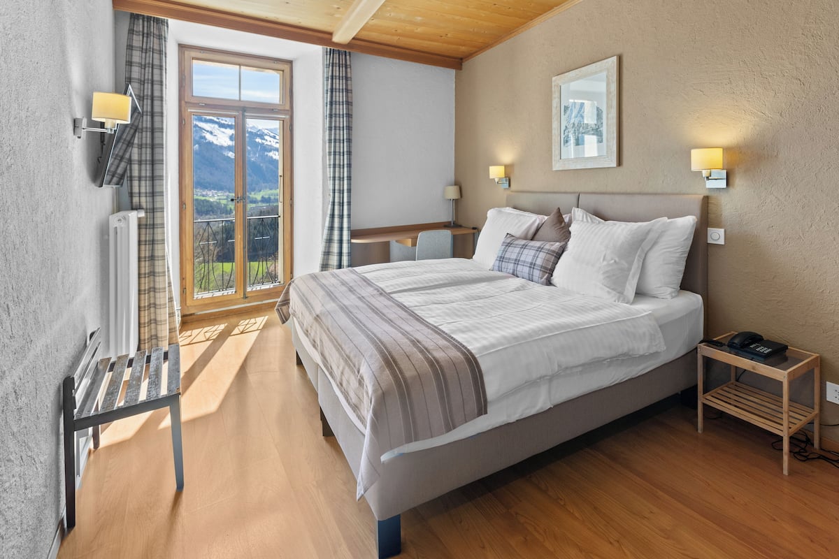 A bedroom with a large bed, a wooden bench, a desk, and a view of mountains through a glass door.