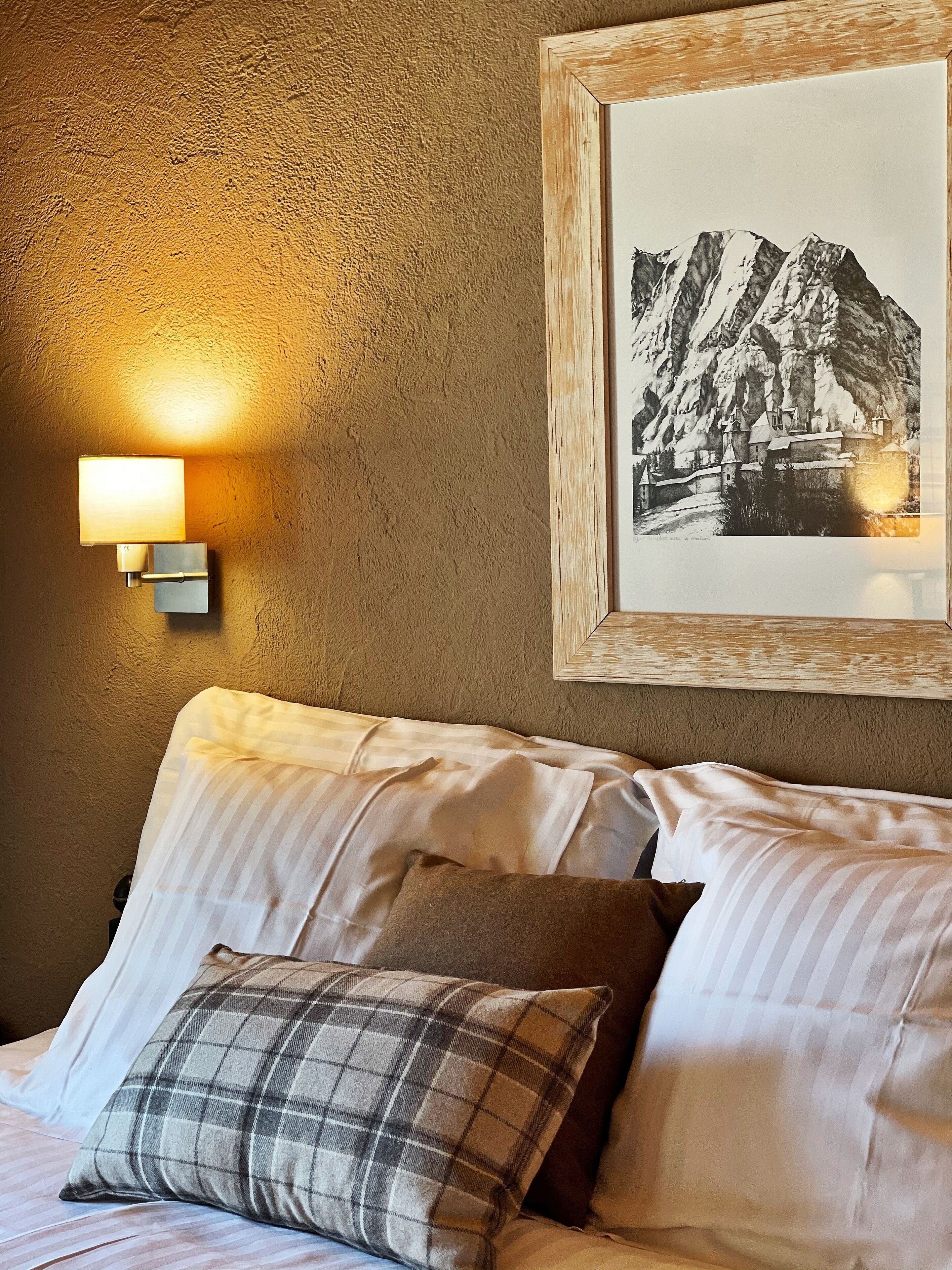 A bed with striped bedding and a plaid pillow, a framed picture of a mountain scene, and a wall-mounted light fixture.