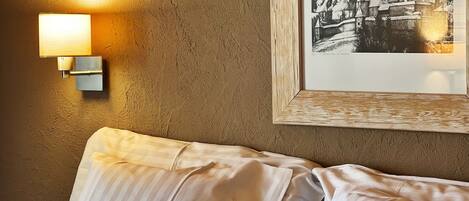 A bed with striped bedding and a plaid pillow, a framed picture of a mountain scene, and a wall-mounted light fixture.