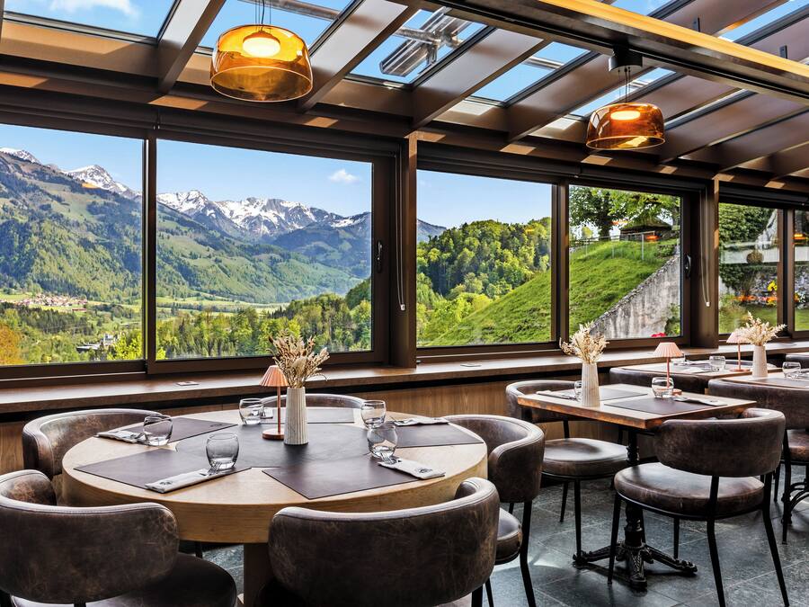 Um restaurante com mesas e cadeiras redondas, janelas amplas com vista para a montanha e luminárias pendentes.