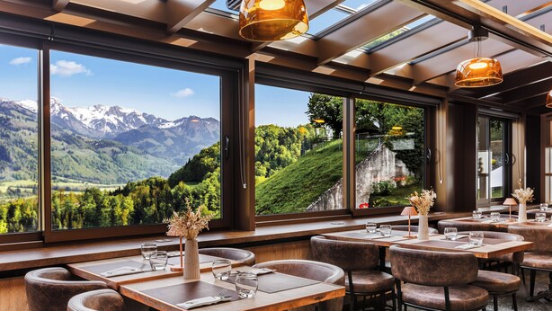 Restaurante com amplas janelas que oferecem vista para as montanhas, mesas de madeira e cadeiras de couro.