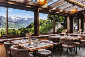 Restaurante com amplas janelas que oferecem vista para as montanhas, mesas de madeira e cadeiras de couro.