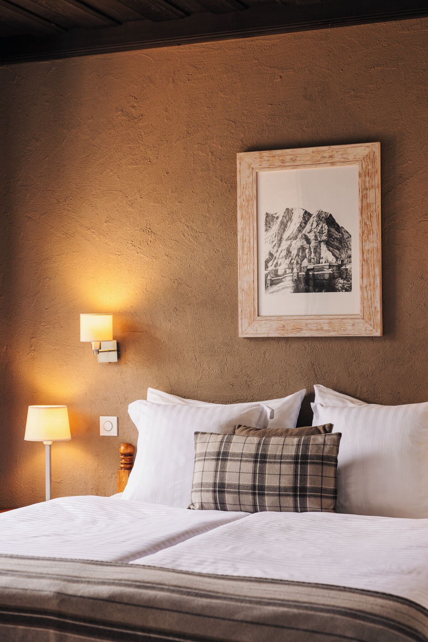 Una cama bien hecha con sábanas blancas y una almohada escocesa, una fotografía blanco y negro enmarcada en la pared, y una lámpara fijada a la pared.