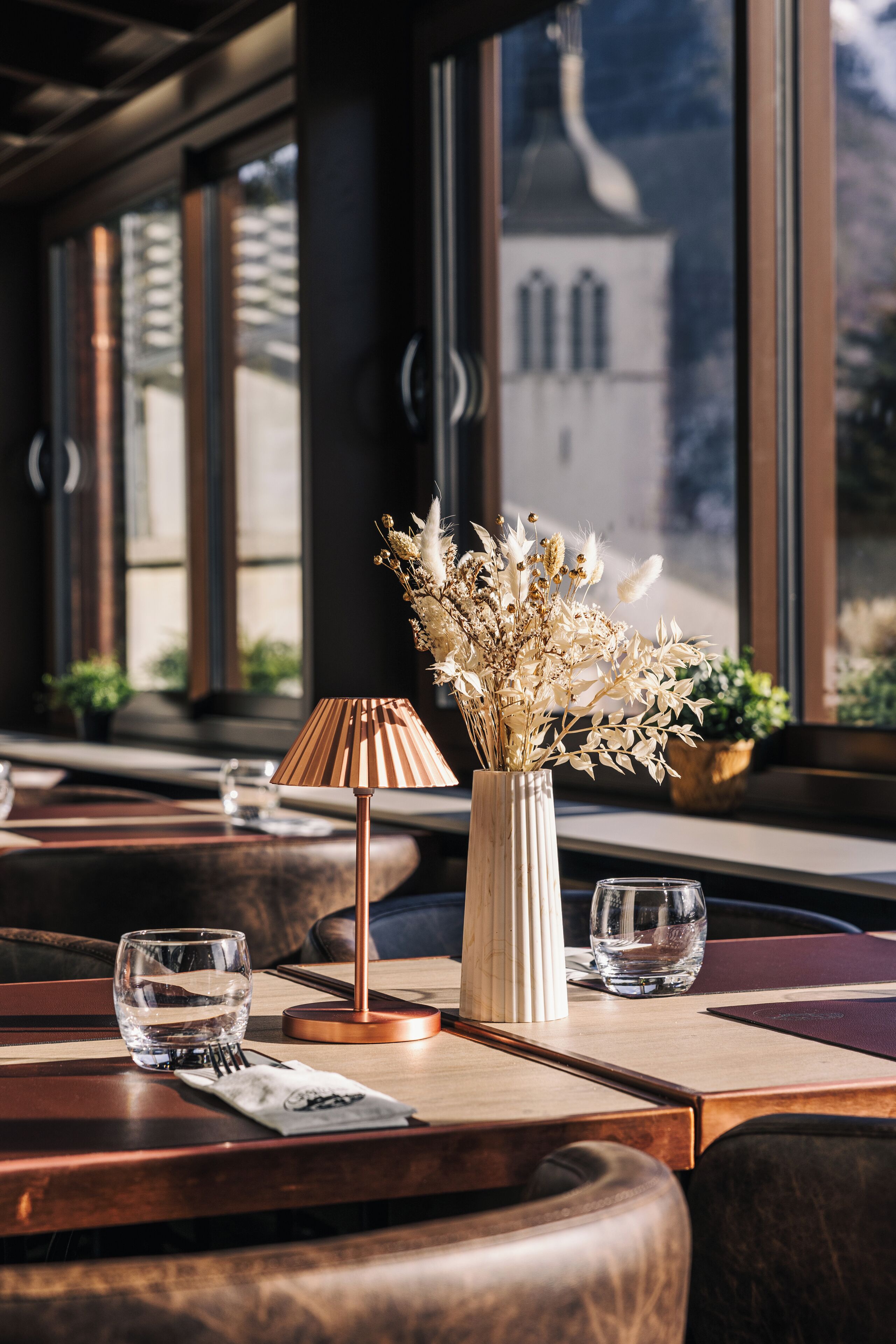 A restaurant interior with a wooden table, a vase with dried flowers, and a lamp.