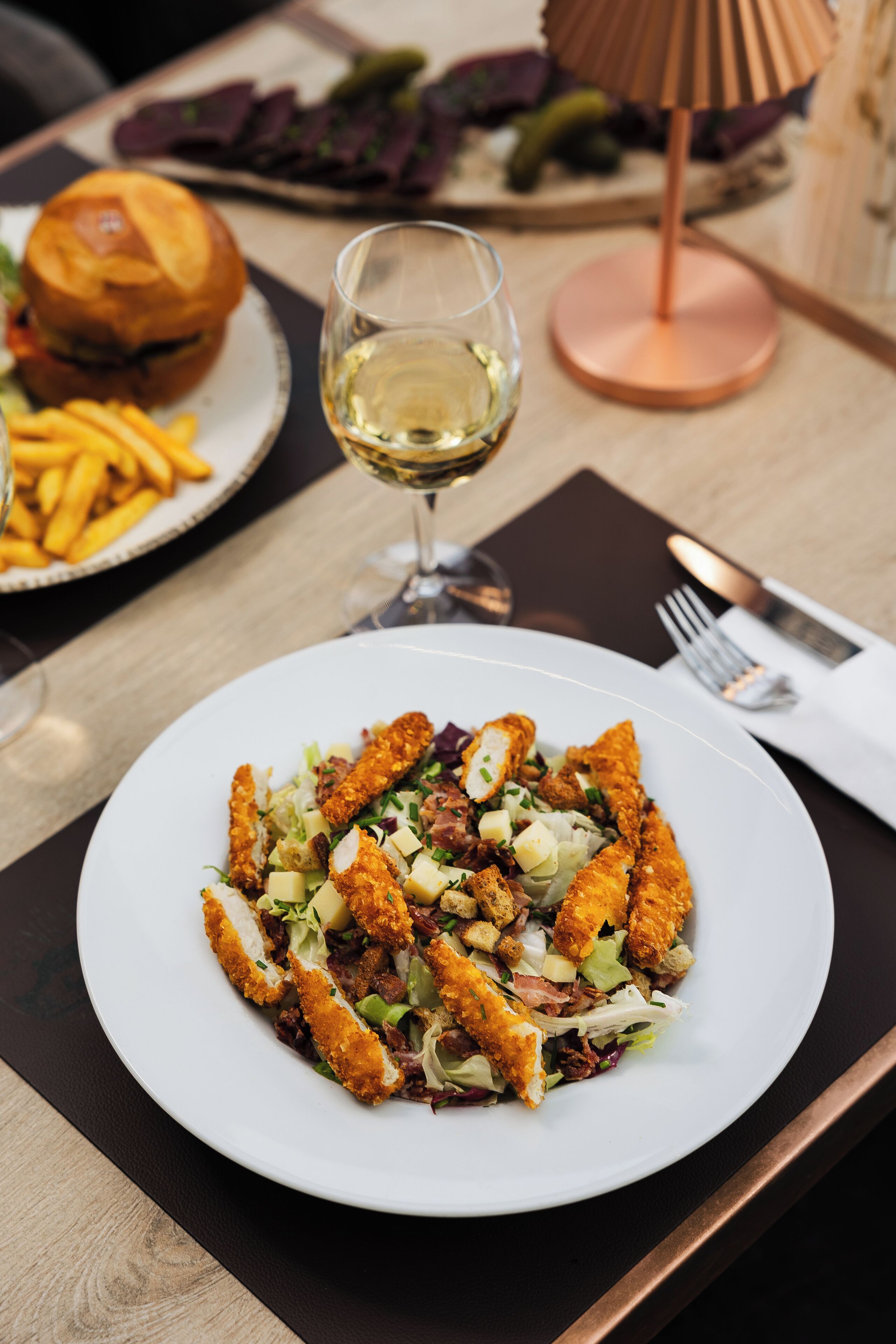 A dining table with a plate of food, a glass of wine, and a burger in the background.