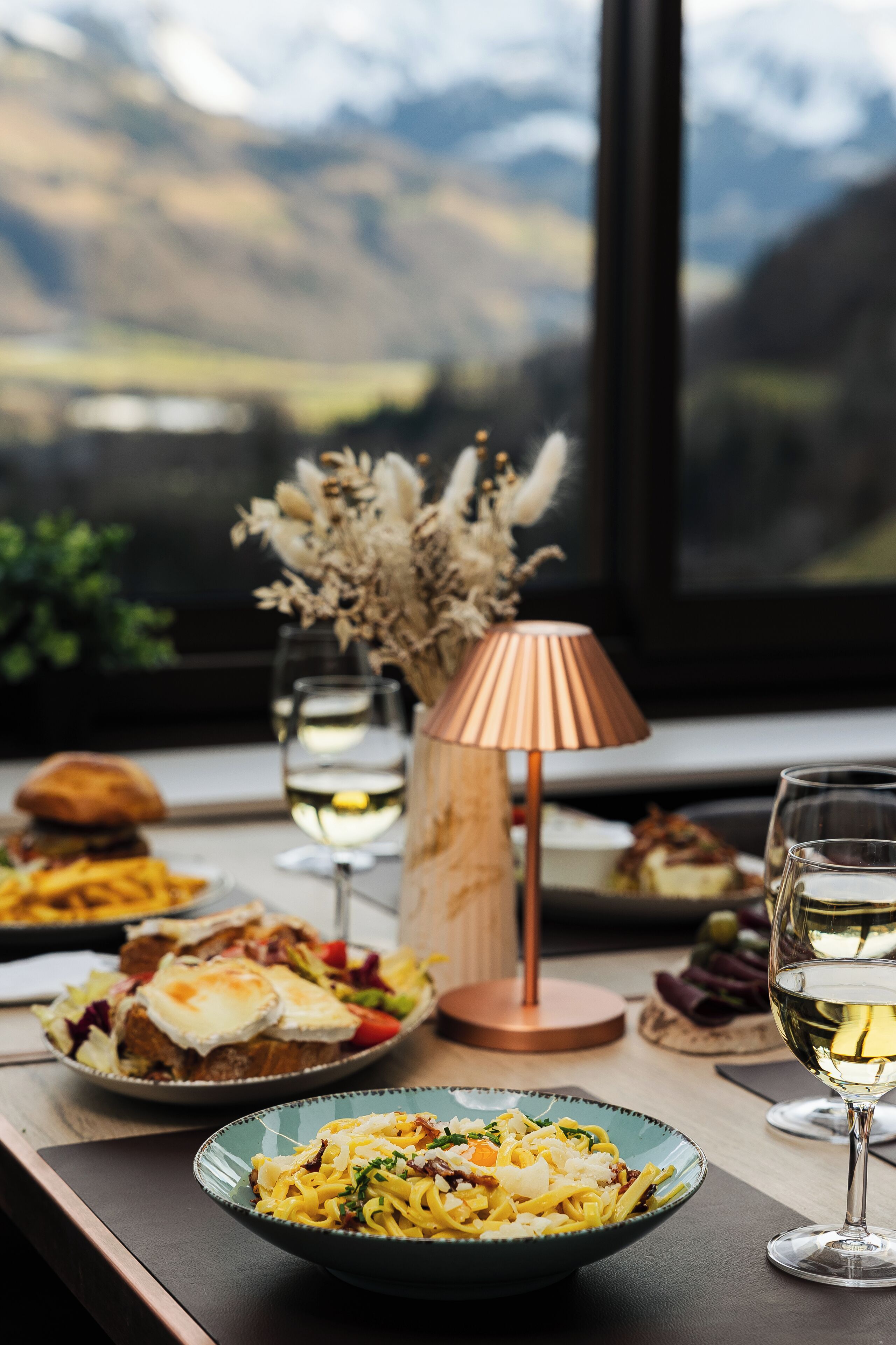 A dining table with various dishes and wine glasses, a lamp, and a view of mountains through a window.