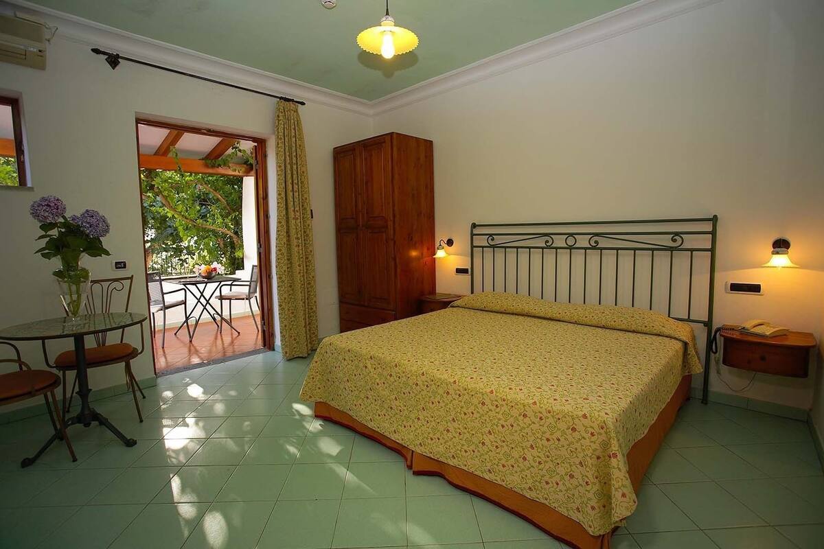 A bedroom with a bed, a table with chairs, a wardrobe, and a balcony with plants.