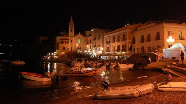 A nighttime harbor scene with boats docked and a building with a tower in the background. - Villa Fiorentino (Lipari)