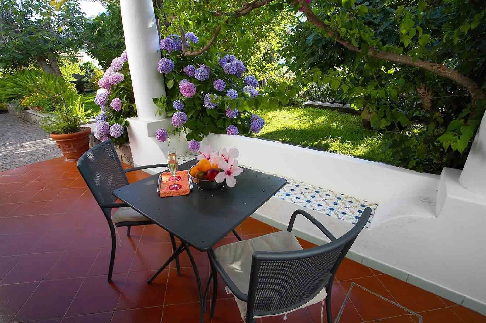 Un patio exterior con una mesa lista para dos personas, rodeado de exuberante follaje y plantas florecientes.