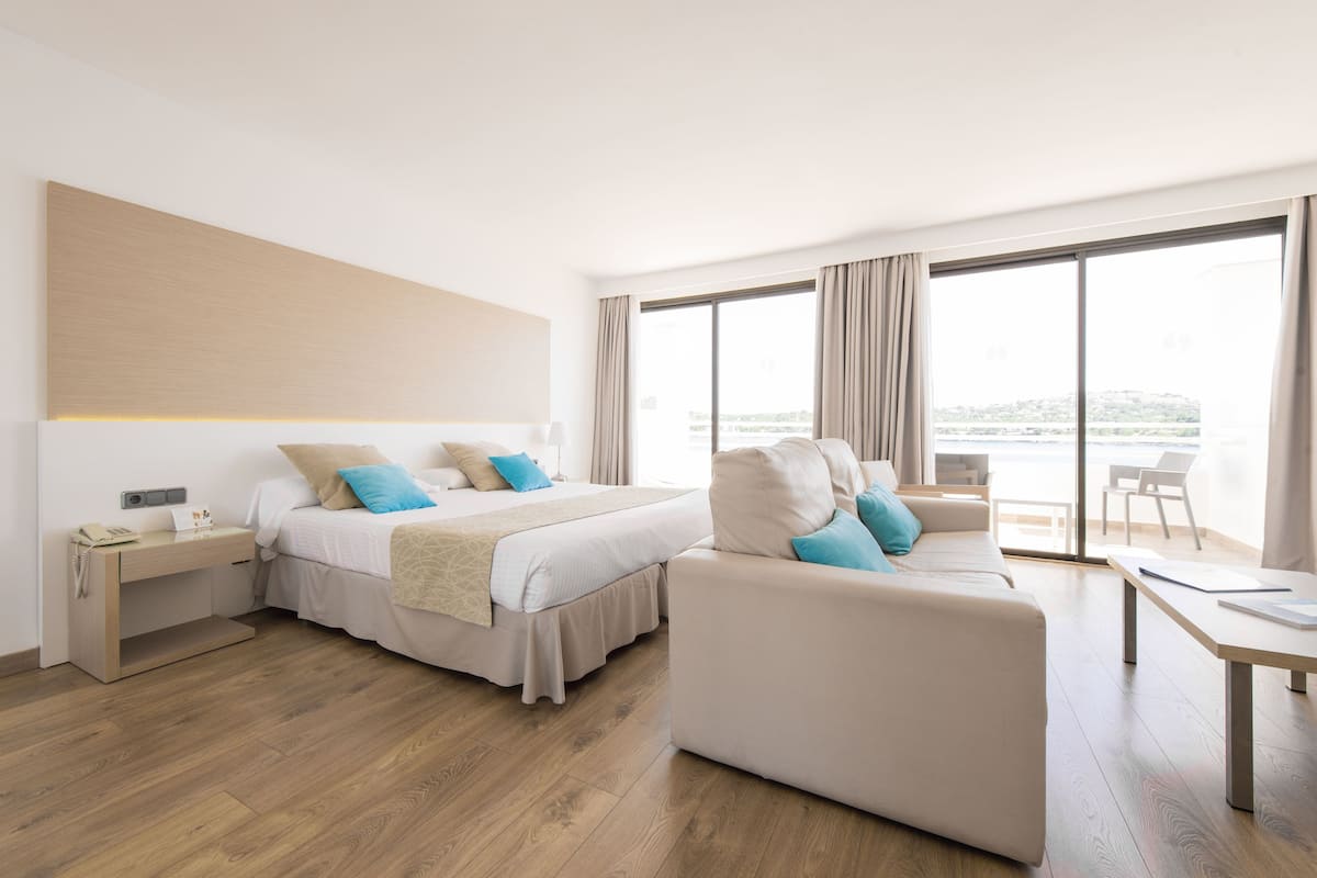 A modern hotel room with a bed, a sofa, a desk, and a large window offering a view.