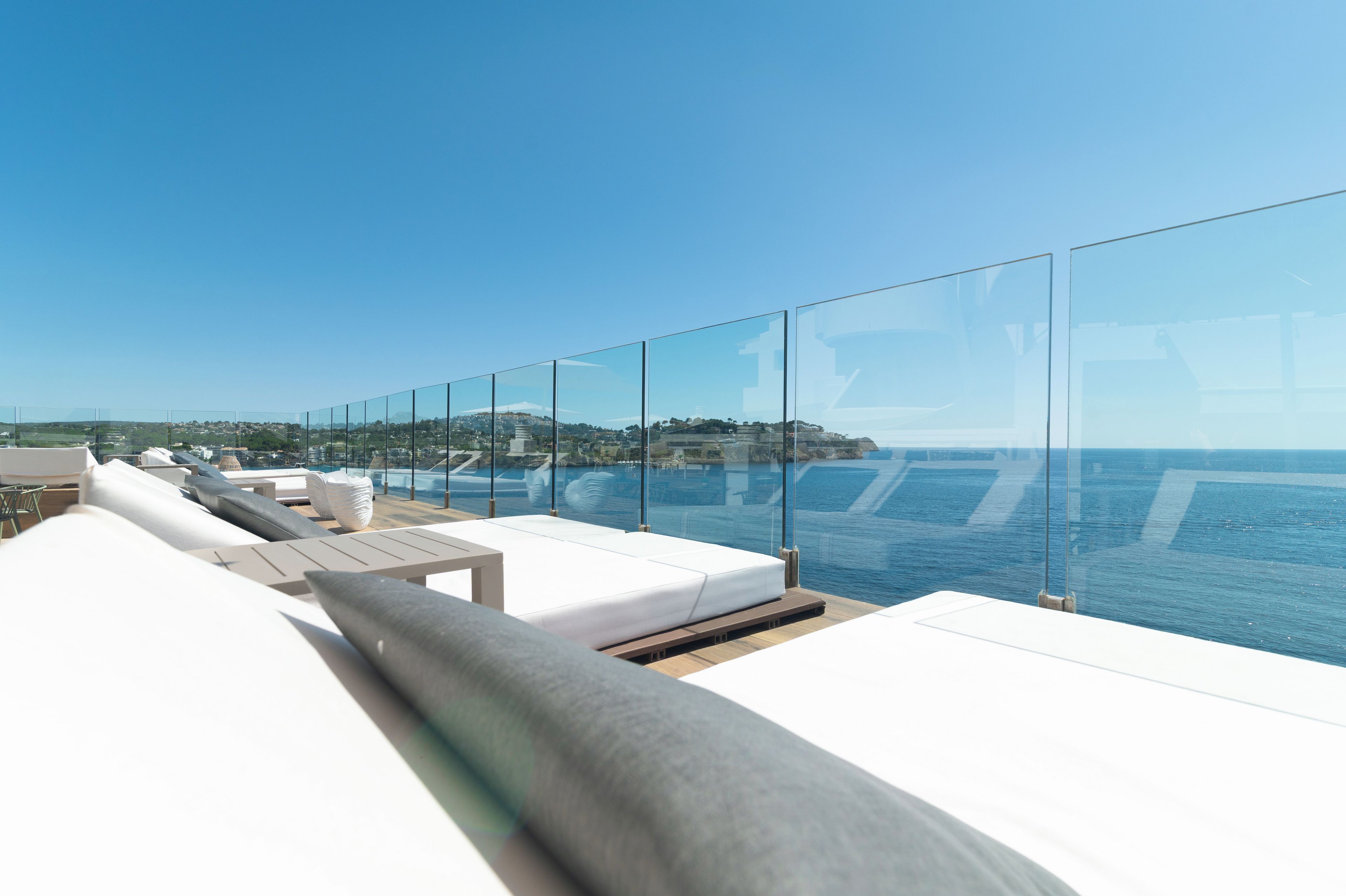 Un salon moderne situé sur un toit-terrasse, doté de sièges blancs et confortables et de rampes de verre, offrant une vue sur une étendue d’eau et sur le paysage urbain lointain.