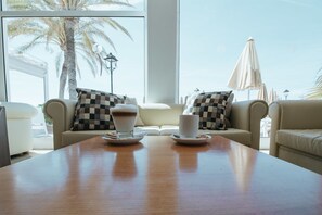 Una habitación con un ventanal grande, dos sofás con cojines de cuadros, una mesa de madera con dos tazas de café y vistas a palmeras y una playa.