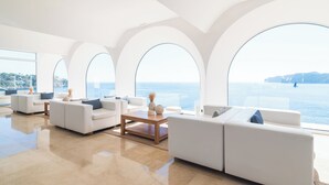Un salón espacioso con sofás blancos, una mesa de centro de madera y grandes ventanales arqueados con vistas al mar.