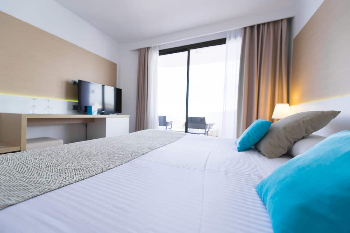 A hotel room with a large bed, a desk with a television, a window with curtains, and a view of the outdoors.