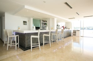 Zona de bar moderna con mostrador de mármol, taburetes blancos y cocina con paredes de vidrio al fondo.