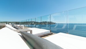 Un moderno salón en la azotea con asientos acolchados blancos y barandas de vidrio con vistas a una masa de agua y un horizonte urbano distante.