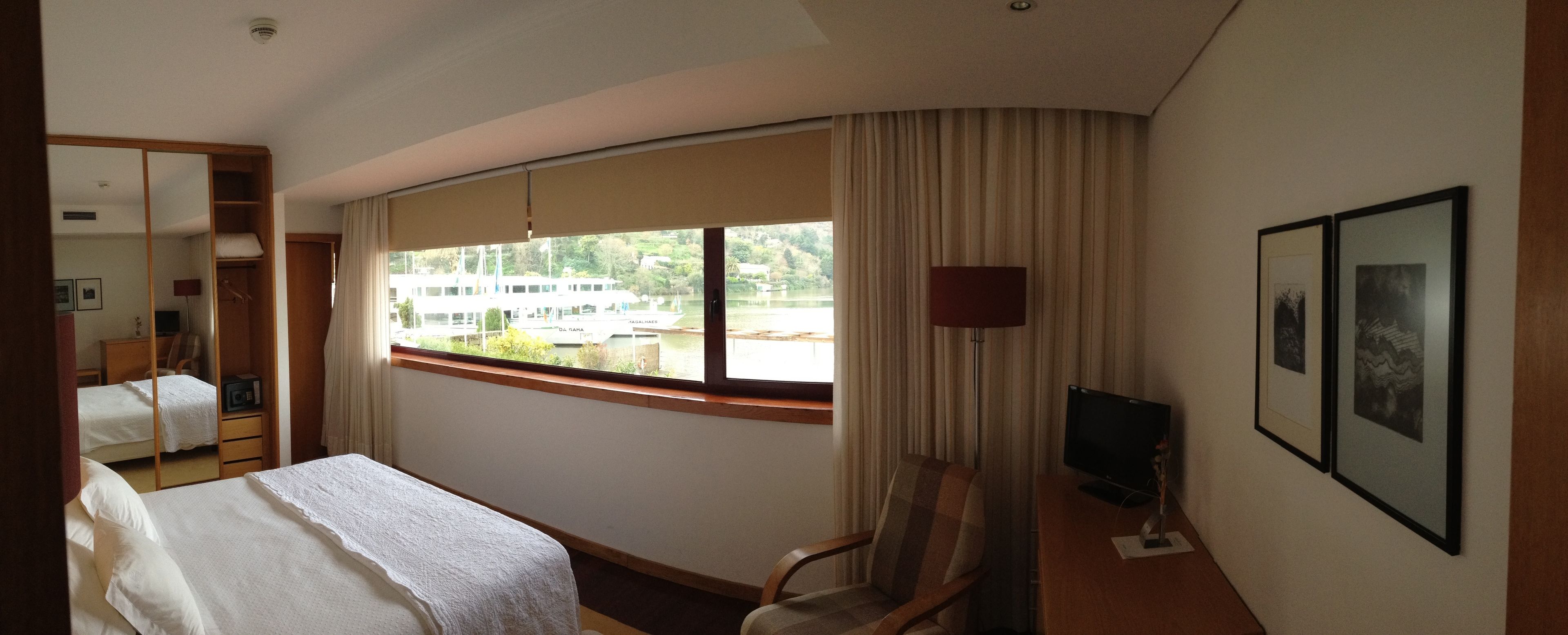 A hotel room with a bed, a chair, a TV, and a window with a view of a boat and greenery.
